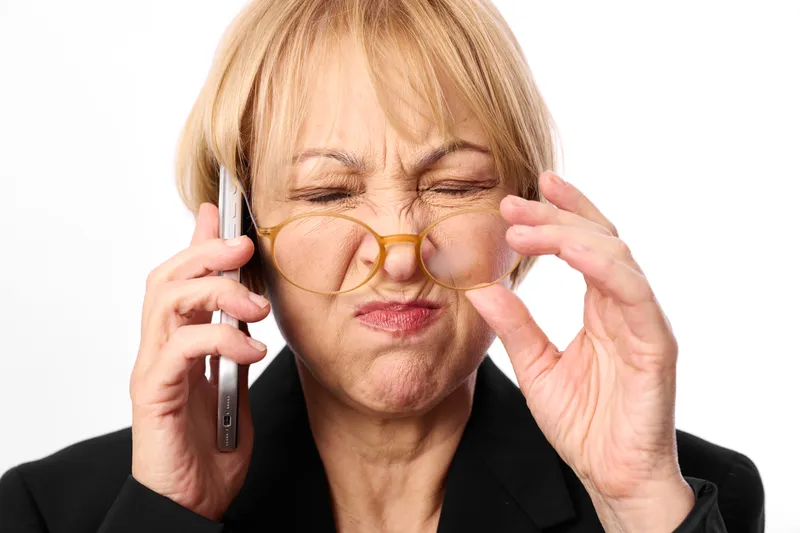 Senior woman speaking on phone call, wearing eyeglasses, adjusting frames with fingers, warm smile, studio portrait, white background, calm communication, soft lighting, closeup