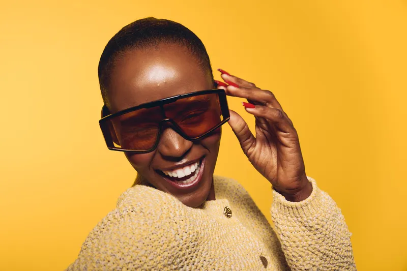 Happy African woman with dark skin wearing oversized sunglasses and a beige knitted sweater smiles joyfully on a yellow background. Concept of fashion, style, confidence, and beauty.