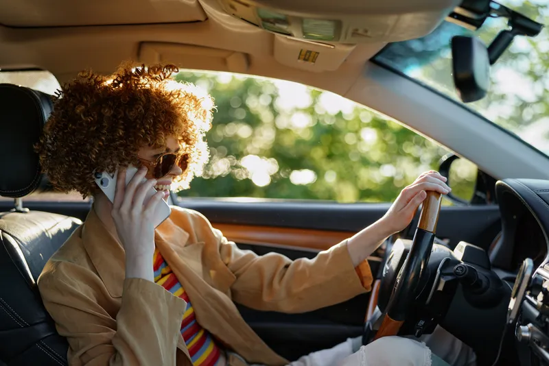 Curly haired driver in beige shirt talks on a cellphone while seated in a car, retro film color and warm lighting create a casual lifestyle moment on the road