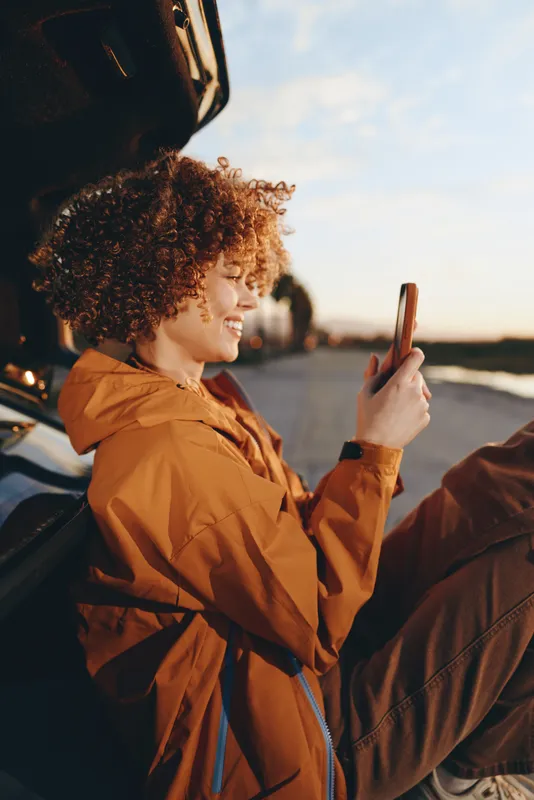 Woman lifestyle smartphone in car, smiling portrait at sunset, curly hair in warm evening light, relaxed travel moment, hands holding phone, road background near window with candid mood