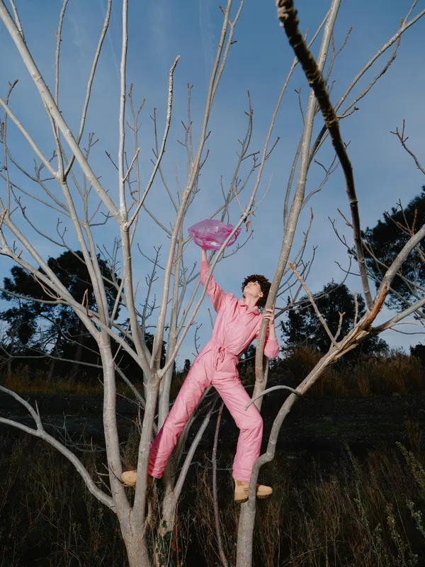 Person in a pink jumpsuit climbs a leafless tree outdoors, reaching with a frisbee in hand, showcasing balance, courage and playful exploration in a natural setting.