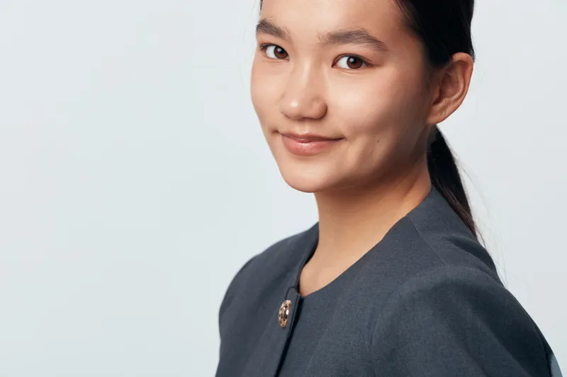 Young woman with dark hair in professional attire posing with slight smile on light background for business portrait or corporate profile photo in studio setting.