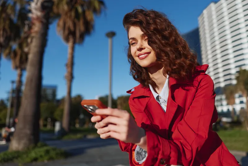 Smiling woman with curly hair wearing a red jacket holding a phone outdoors on a sunny day, modern buildings in the background, cheerful and casual vibe.
