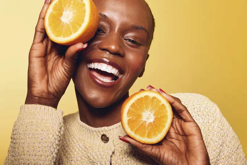 Smiling dark skinned African woman holding two orange halves near her face on yellow background. Concept of healthy lifestyle, freshness, vitamin C, natural beauty, and skincare.