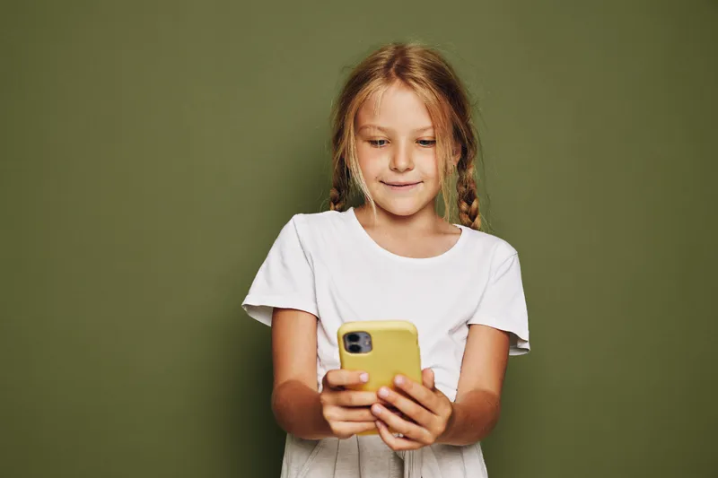 Smiling girl with braided hair wearing white t-shirt looking happily at her yellow smartphone on green background.