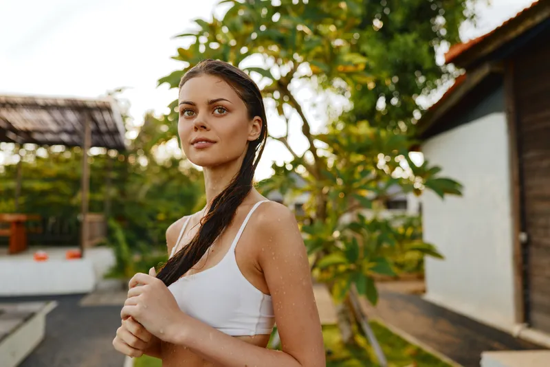 A lifestyle portrait of a woman outdoors in a sunny backyard, wearing a white tank top, hair in a braid, posing casual and relaxed in natural light.