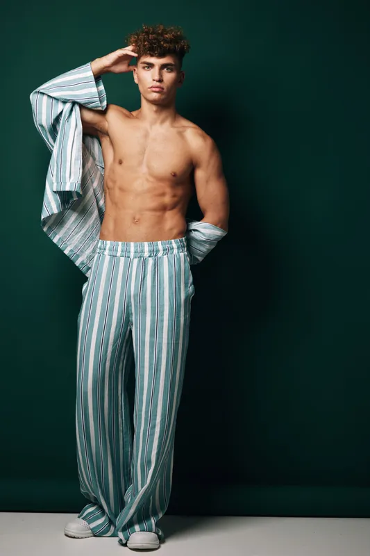 Young man with curly hair posing shirtless wearing striped pants and partially removed shirt against dark green background in studio. Fashion model with casual style and confident expression.