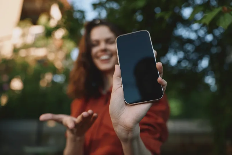 Bright outdoor moment with a smiling woman showing her phone, casual lifestyle, natural light, vibrant greenery in the background, candid and relaxed, soft breeze