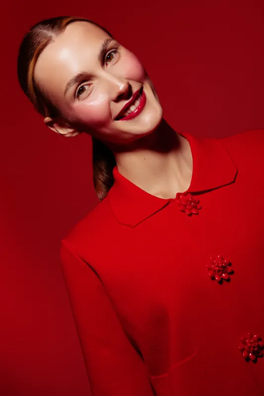 Smiling woman with red lipstick and red coat poses against red background. Studio portrait with bright lighting highlights her clear skin and confident expression. Concept of fashion and beauty.