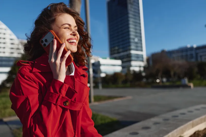 A young woman in a red coat talks on her smartphone outside a modern cityscape, smiling brightly as she enjoys a sunny day and the urban breeze around her.