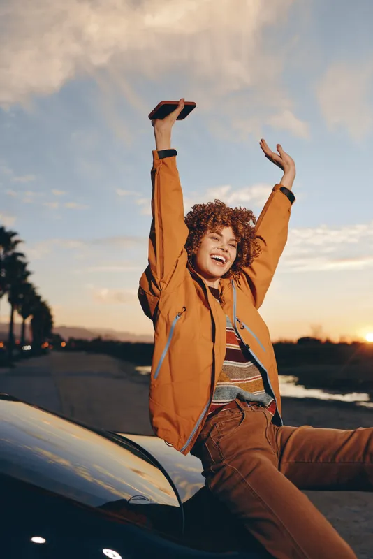 Woman lifestyle outdoors sunset car jacket joyful laughing, curly haired woman on car hood reaching up with arms, warm evening light, relaxed pose, road background, natural colors