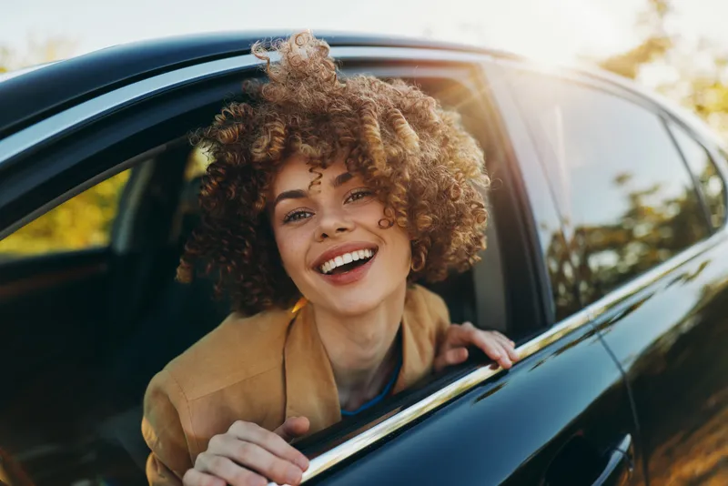 Joyful young woman with curly hair leaning out of a car window smiles brightly in warm sunlight, capturing a spontaneous carefree moment.