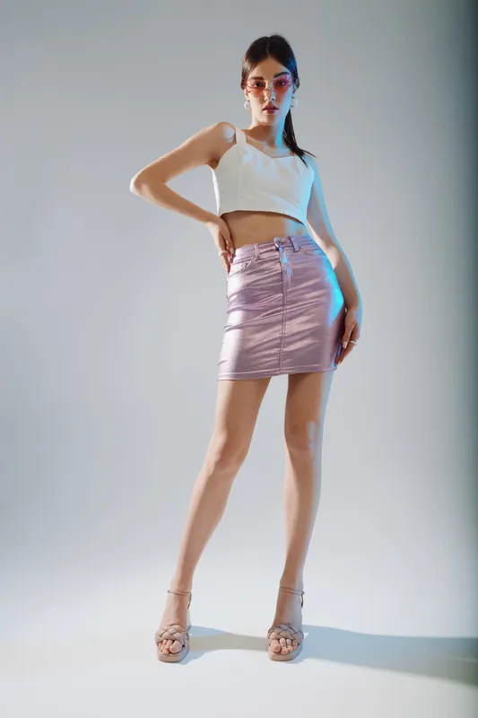 Young woman in a white crop top and purple skirt poses confidently in high heels. Studio shot with soft lighting on a neutral background. Concept for woman beauty fashion style.