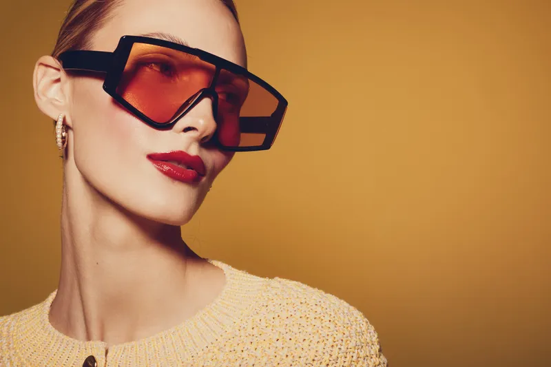 Stylish woman with oversized orange sunglasses and red lipstick poses against a warm yellow background. Fashion concept with modern accessories and confident expression.