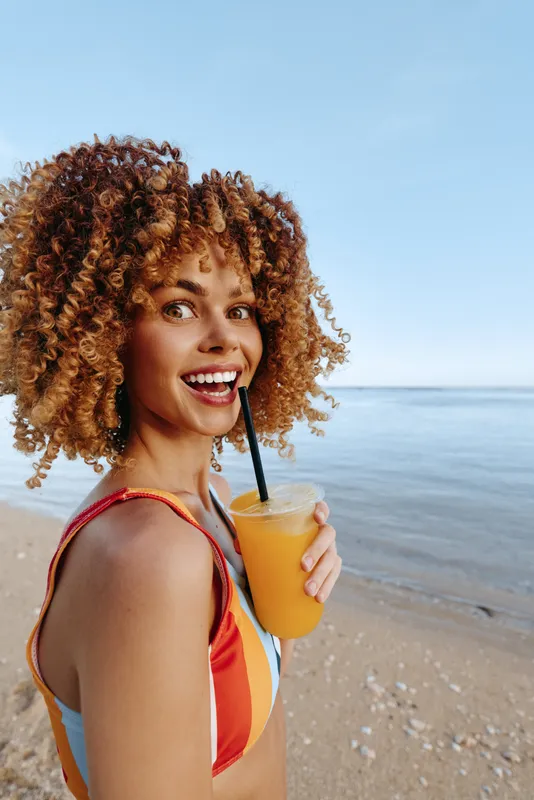 Woman with curly hair on beach, ocean sunlight background, smiling summer vacation, holding a juice drink, outdoors on sand, seaside relaxation, natural portrait