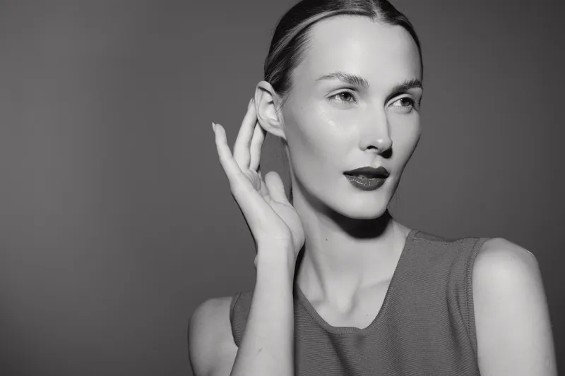 Elegant Caucasian woman with fair skin and slicked back hair poses with hand near ear against plain background, showcasing beauty and grace.