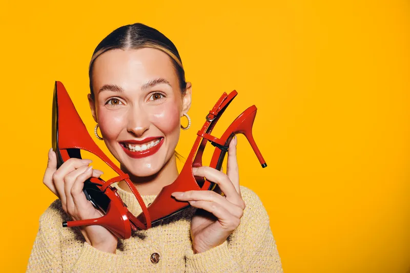 Smiling woman with red lipstick holds red high heel shoes near face on bright yellow background. Concept of fashion, style, footwear, and joyful expression in studio lighting.
