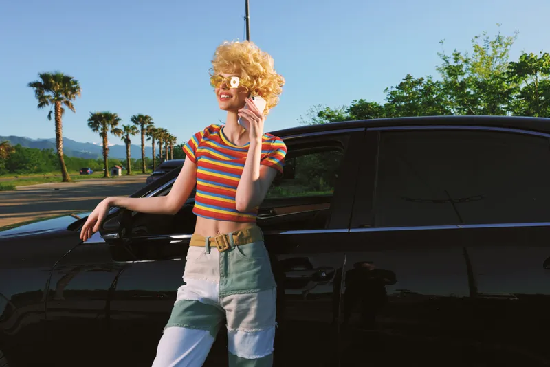 Woman lifestyle shot with retro color tones leans on a black car, wearing a striped crop top and patchwork jeans, cheerful pose and sunny outdoor vibe. Her confident smile and stylish look evoke a