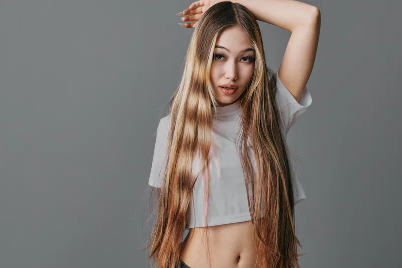 Young woman with long straight hair wearing a white crop top posing with hand raised in studio on gray background. Beauty, fashion, style and confident female portrait.
