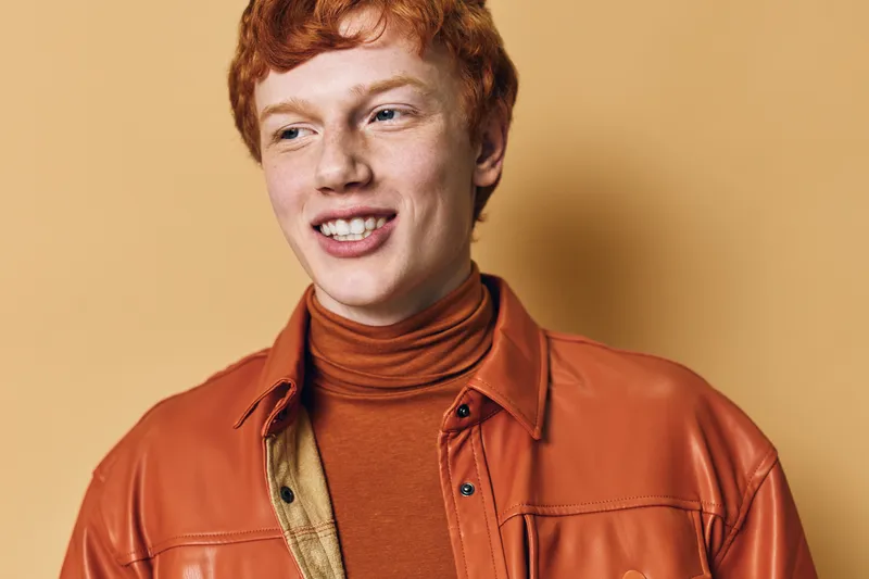 Young man with red hair and fair skin smiling in leather jacket and turtleneck against neutral background. Casual portrait with natural light and confident expression.