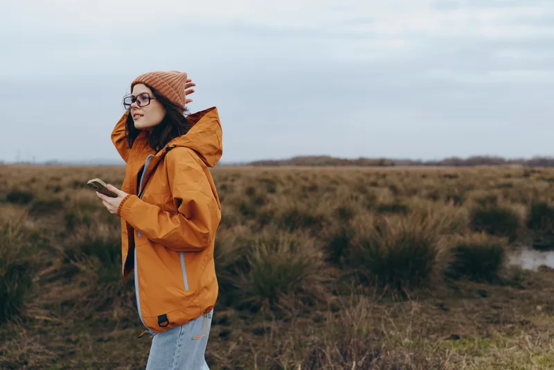 Outdoor nature lifestyle portrait of a woman in orange jacket, hat and glasses, holding a smartphone as wind lifts her hair in an autumn grassy field under cloudy sky