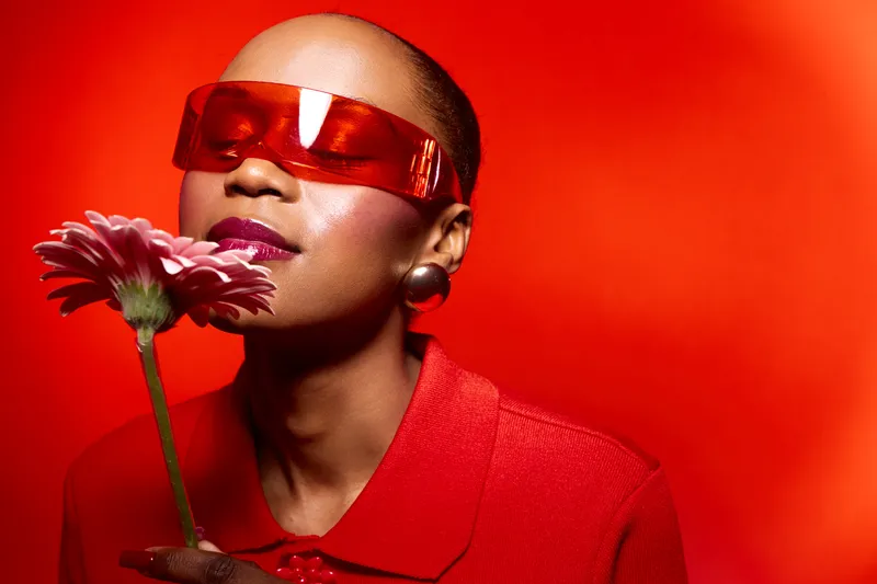 Confident black woman with dark skin wearing red sunglasses and red outfit holds pink flower close to face on vibrant red background. Concept of fashion, beauty, and elegance in studio.