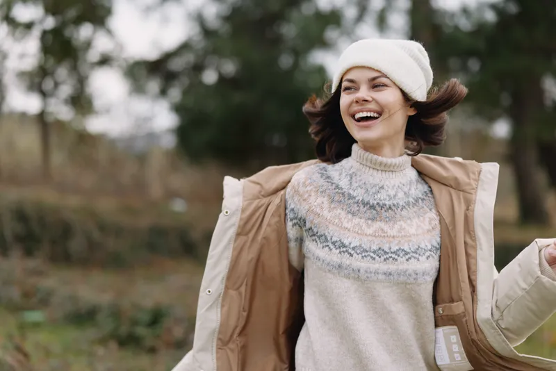 Woman outdoors wearing a knit sweater and coat, smiling joyfully on a windy day, fashionable autumn style with a hat, open countryside background.