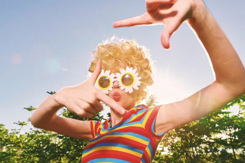 A retro lifestyle outdoors scene captures a playful, colorful portrait of a person wearing sunglasses in bright daylight, conveying whimsy and bold fashion in a sunny, candid moment.