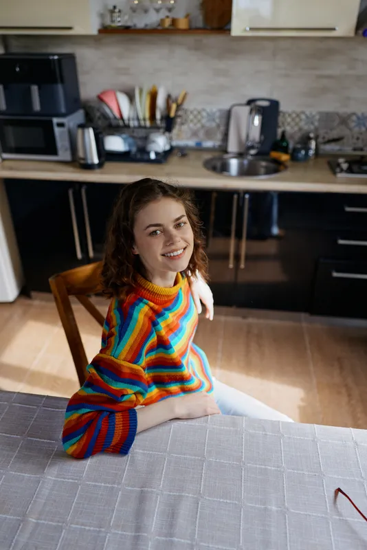 A smiling woman wearing a vibrant striped sweater sits at a kitchen island, casual and cozy at home. Natural light highlights her relaxed mood and everyday lifestyle.