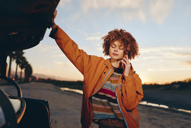 Woman lifestyle outdoors at sunset beside a car, wearing a casual jacket, curly hair in the breeze, relaxed smile, standing by open door, warm light over road