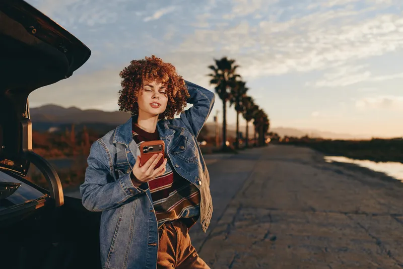 Woman lifestyle portrait outdoors at sunset, holding phone near car with open trunk, wearing denim jacket and orange dress, curly hair, palm lined road, golden hour mood calm.