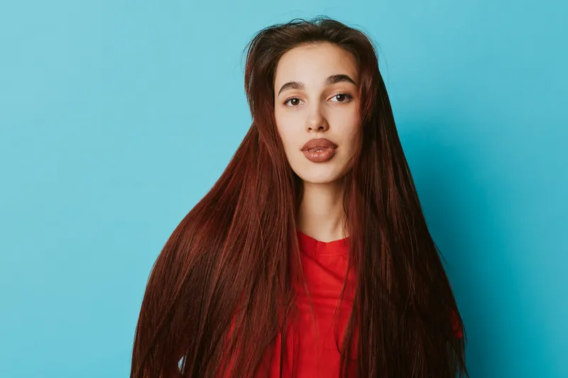 Young woman with long straight hair wearing red shirt posing against blue background looking at camera with neutral expression in studio portrait with simple colorful backdrop.