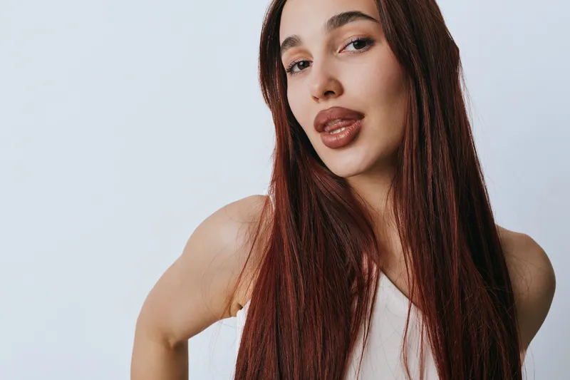 Woman with long brown hair and natural makeup posing confidently in white top against light background. Portrait of young female with smooth skin and subtle expression.