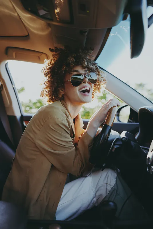 A woman driver in a car embodies lifestyle and retro vintage vibes, wearing sunglasses as she smiles. A candid road trip scene in warm film color tones and joyful energy.