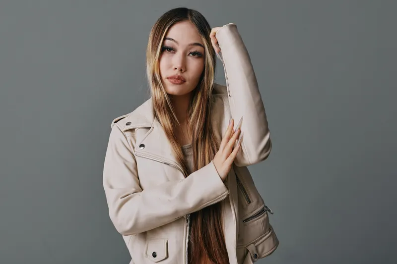 Young woman posing in beige leather jacket with long straight hair against grey background, looking confident and stylish in casual fashion portrait.