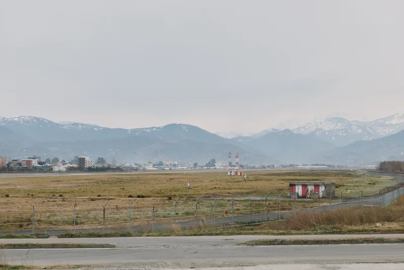 Empty rural landscape with overcast sky and distant mountains, featuring grassy fields and small buildings near a paved road. Concept of countryside, nature, and calm environment.