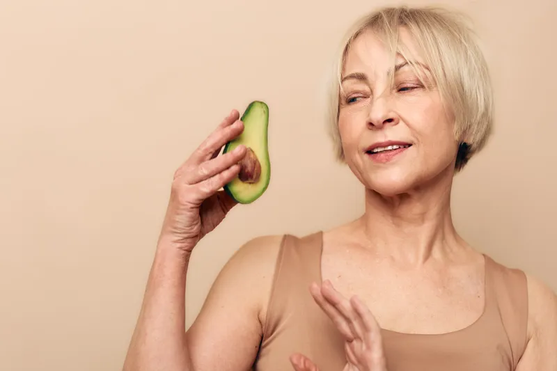 Mature woman with short blonde hair holds half avocado near face, smiling gently with closed eyes. Concept of healthy lifestyle, natural beauty, nutrition, and wellness for seniors.