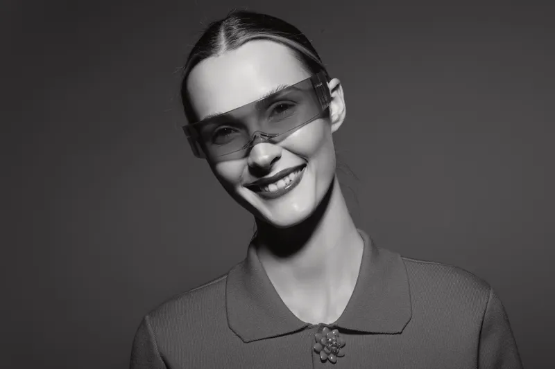 Smiling Caucasian woman with light skin wearing futuristic sunglasses and vintage outfit looks confident in studio portrait with dark background.
