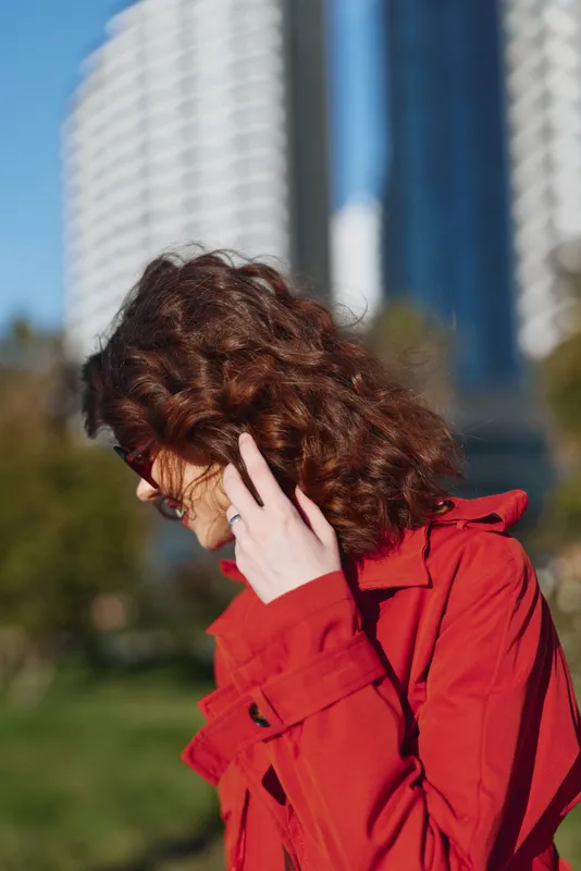 Curly-haired woman in a red coat adjusts her glasses outdoors, with a bright city backdrop. Fashion portrait capturing style, confidence, and urban elegance.