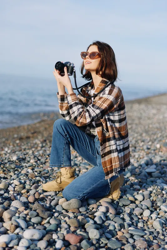 Woman photographer on a rocky beach, kneeling with a camera and lens, wearing a plaid shirt, jeans and sunglasses, capturing coastal scenery under a clear sky for creative outdoor imagery.