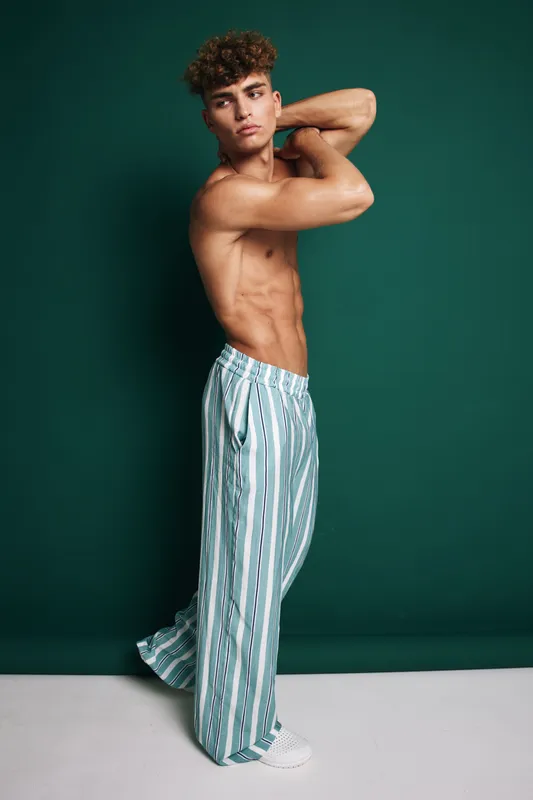 Young man with muscular body posing shirtless in striped wide pants and white sneakers against green background. Fashion, fitness and casual style with confident expression and curly hair.