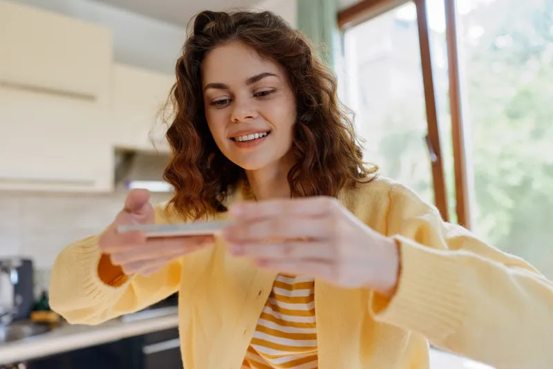 Cheerful woman at home looks at her smartphone, wearing a soft yellow cardigan, bright kitchen in the background, casual lifestyle moment with relaxed warm mood and everyday connectivity