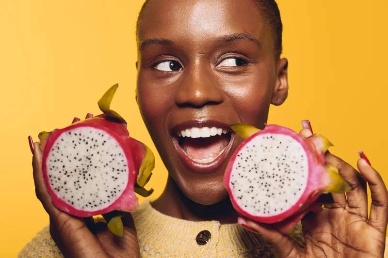 Smiling African woman with dark skin holds two halves of a dragon fruit near her face. Bright yellow background highlights fresh tropical fruit and joyful healthy lifestyle concept.