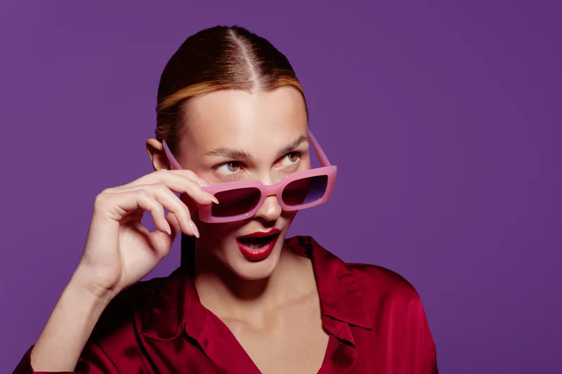 Young woman in red shirt and pink sunglasses looks surprised against purple background. Fashion portrait with trendy accessories and expressive facial expression.
