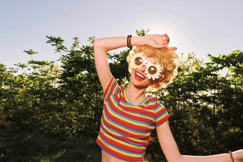 A young woman in a colorful striped top wears a flower mask and playful pose outdoors, capturing a carefree mood and bold, whimsical style.
