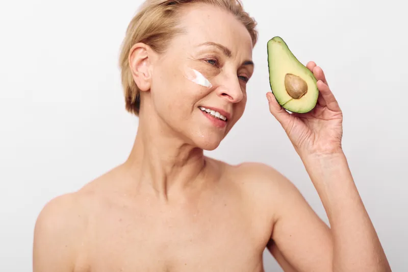 Smiling mature woman with short hair applying cream on face holds fresh avocado half near head. Concept of natural skincare, healthy lifestyle, beauty routine, and organic cosmetic care.