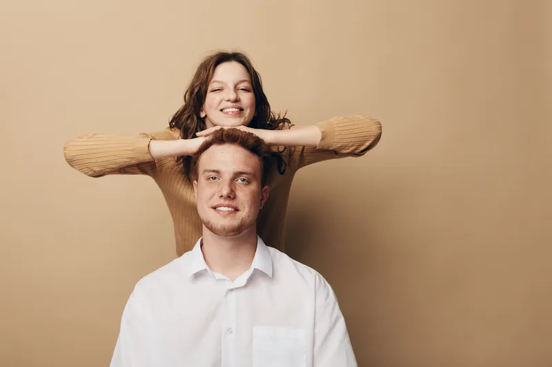 Smiling young woman in brown sweater rests hands on head of seated man in white shirt. Both look relaxed and happy against beige background. Concept of friendship, casual style, and positive mood.