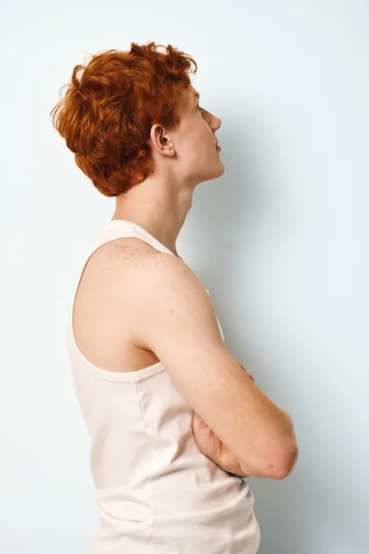 Young redhead man with freckled skin in white tank top standing against white wall with crossed arms, looking sideways in casual pose and natural light indoor portrait.