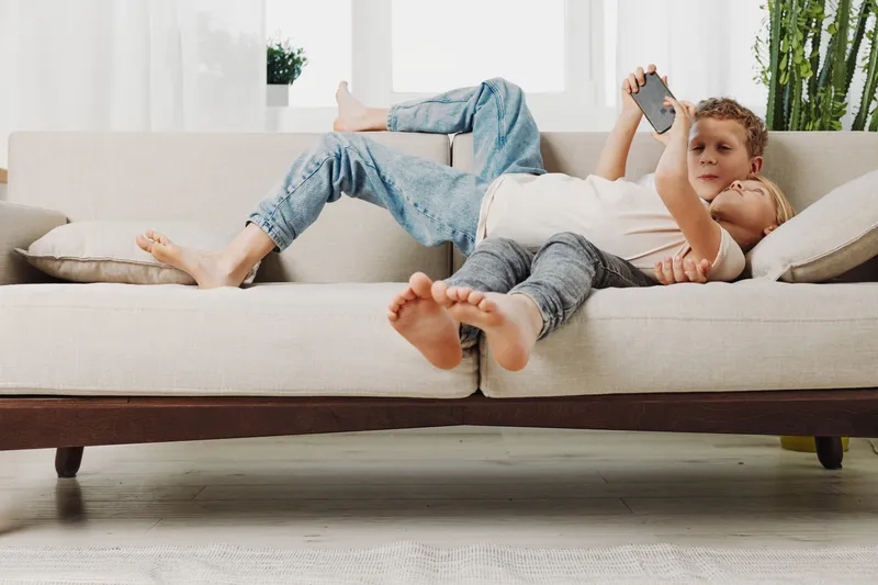 Two young boys lying on a beige sofa at home, one holding a smartphone while the other rests, natural light brightens the modern living room with plants and cushions.