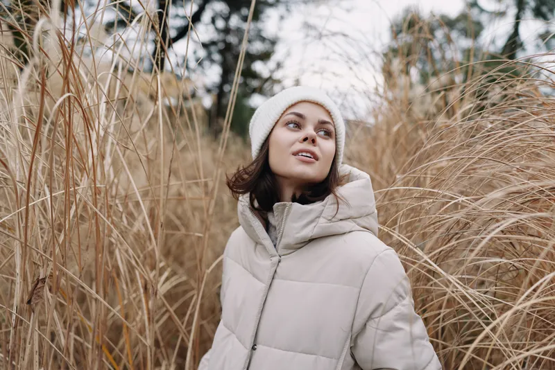Woman in a light winter coat stands among tall dry grass, looking up with a soft smile in an open field, chilly air and natural light creating a calm seasonal portrait for lifestyle and fashion use.